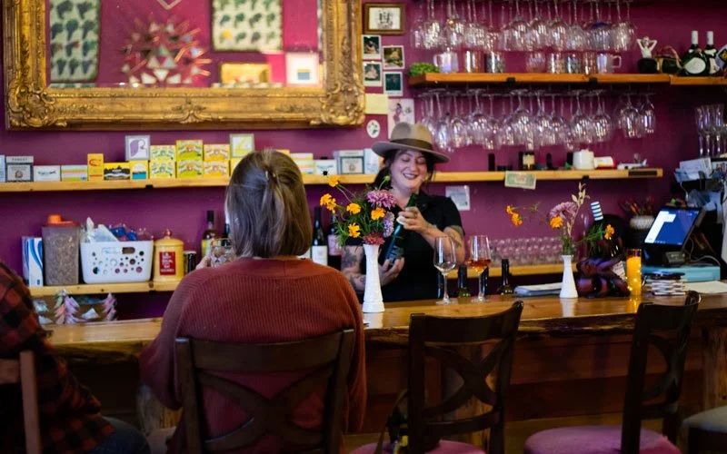 A woman with a wide-brimmed hat and black shirt serving drinks behind a bar, with two patrons seated in front, in a cozy, colorful cafe or wine bar with glassware and floral arrangements.