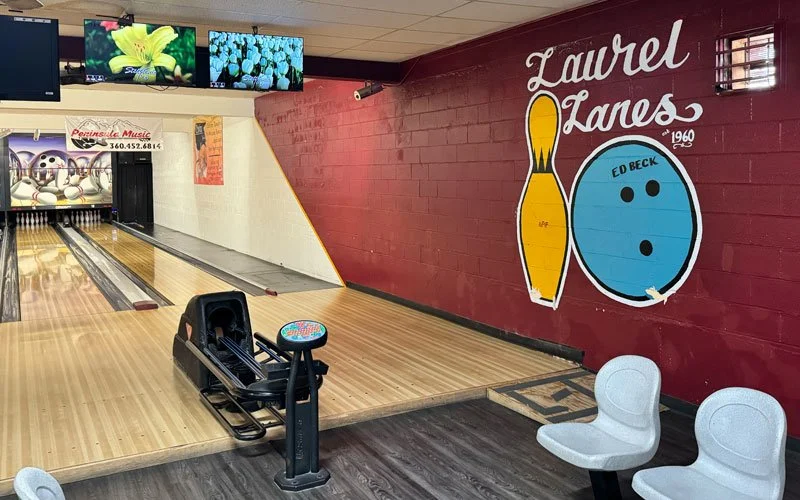 Bowling alley with wooden lanes, bowling balls, scoring screens, a mural with a bowling pin and ball, and seating area.