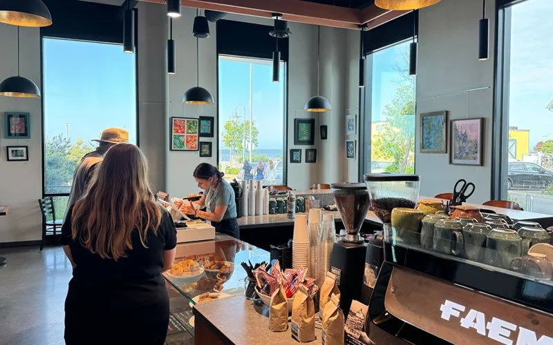 Interior of a coffee shop with large windows, artwork on walls, and customers waiting at the counter.