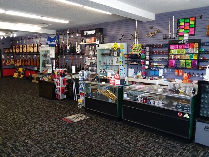 View inside a music store with guitars hanging on the wall to the left, various instrument accessories and music supplies on shelves and counters, including a Yamaha display, and a display of colorful sheet music or accessories on the right.