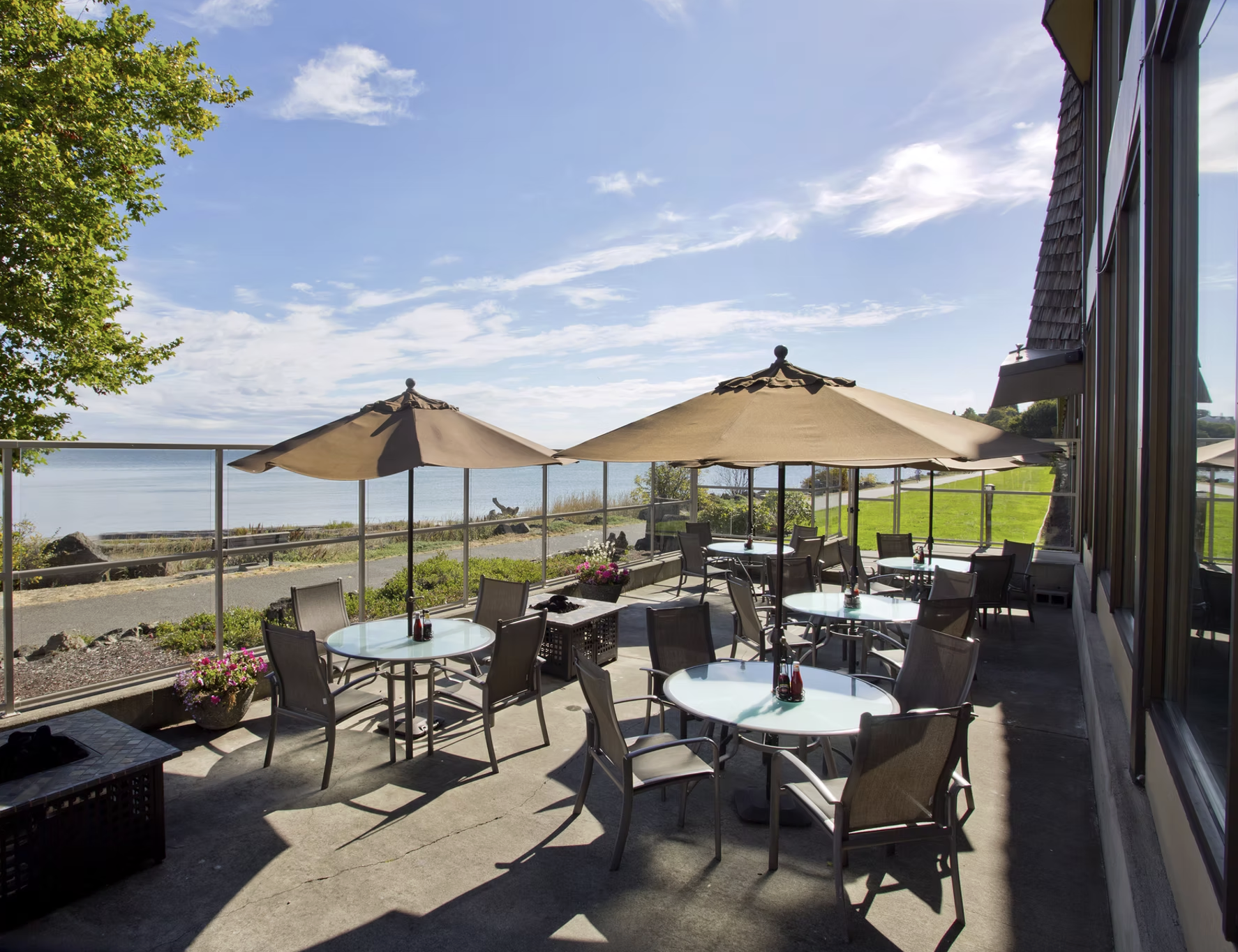 Outdoor patio with round tables, chairs, and large umbrellas overlooking a lake under a partly cloudy sky.