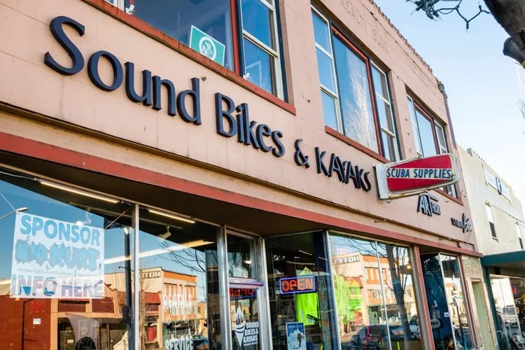 Exterior view of a store named 'Sound Bikes & Kayaks' with a sign for 'Scuba Supplies' on the upper right. The store has large windows and an 'Open' sign in the window.