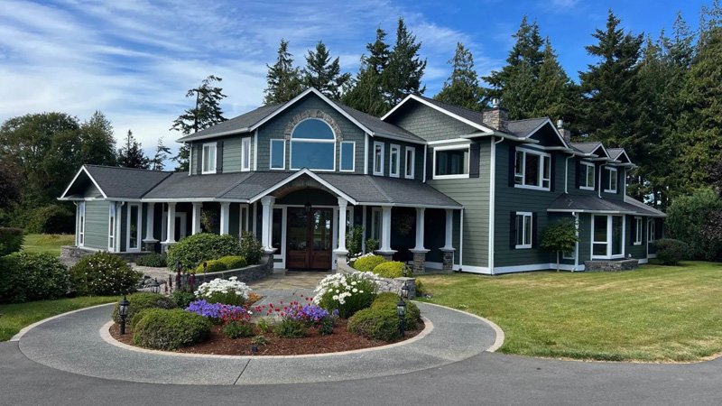 Large gray and green house with multiple stories, surrounded by a well-maintained lawn and flower beds, set against a backdrop of tall trees and a blue sky.