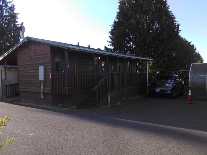 A small wooden building with steps leading up to a porch, situated next to a parking lot with a black vehicle and a traffic cone, surrounded by trees.