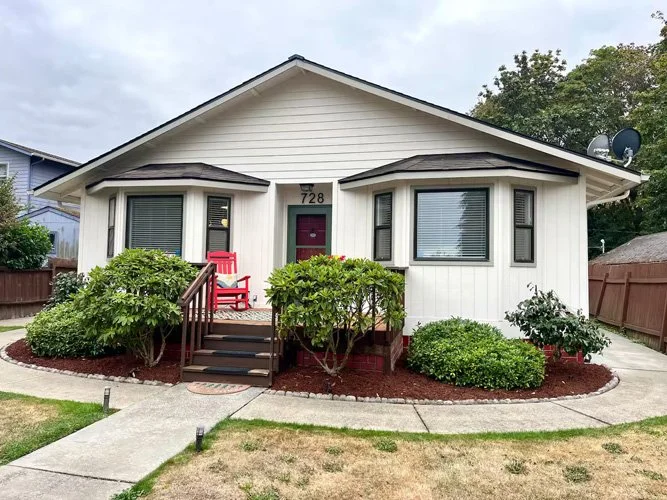 Front view of a white house with a gable roof and a red door, decorated with a red Adirondack chair on the porch, surrounded by landscaped bushes, with a concrete walkway leading to the porch.