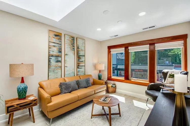 Living room with tan sofa, wooden side table, and large window with wooden trim.