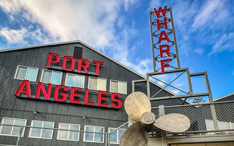 View of the Port of Los Angeles building with large red signs saying "PORT" and "ANGELES" and a vertical sign that reads "WHALE" on a metal frame, a boat propeller, and a blue sky with some clouds.