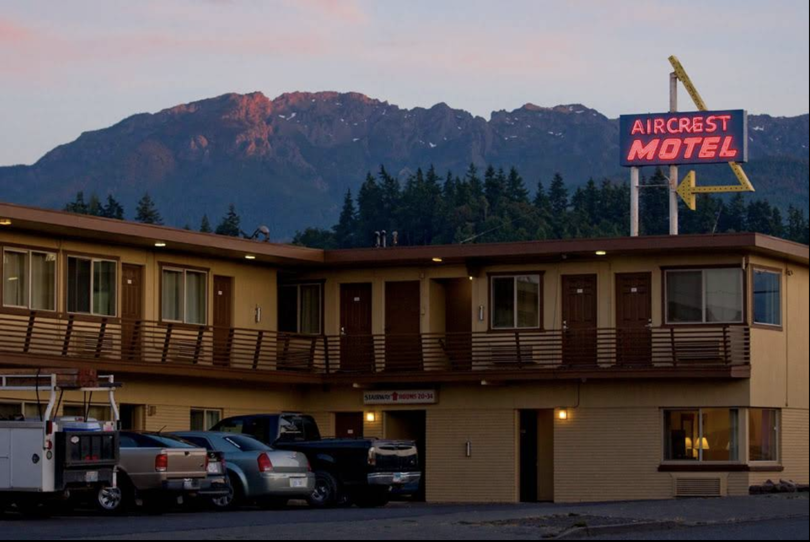 A two-story motel building with a sign that reads 'Aircrest Motel' and a neon arrow pointing left, set against a backdrop of mountains and trees at dusk. There are cars parked in front of the motel.