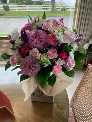 A large bouquet of pink, purple, and white flowers, including roses and carnations, in a white paper wrapper on a table near a window.