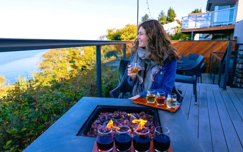 A woman smiling and holding a glass of wine on a balcony deck overlooking a body of water and trees, with a portable fire pit and a tray of six glasses filled with different colored drinks on the table.