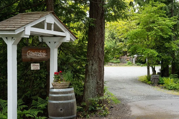 Entrance to a winery called Camadachie, with a wooden sign indicating it is open, surrounded by trees and greenery, with a gravel driveway leading into the property.