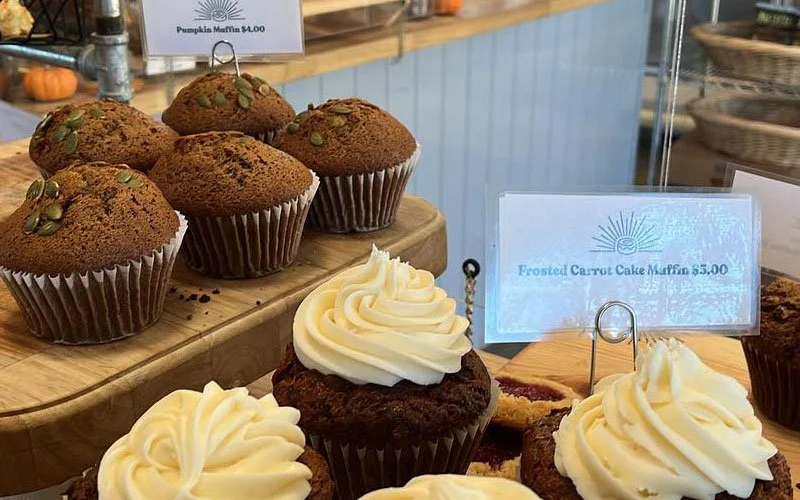 Display of pumpkin muffins and frosted carrot cake muffins at a bakery, with signs showing prices.
