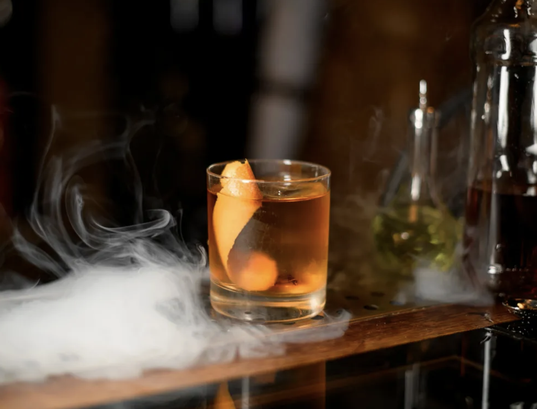A glass of whiskey with an orange peel in it, sits on a wooden table top with smoke surrounding it.