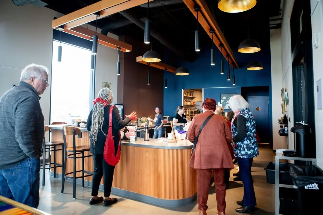 Interior of a coffee shop with large windows, artwork on walls, and customers waiting at the counter.