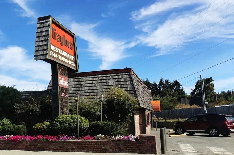 Exterior of Traylor's restaurant with a large sign, brick building with shingle roof, surrounded by greenery and parking lot with black and red cars, under a blue sky with some clouds.