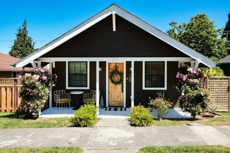 Small black house with white trim, decorated for Christmas with a wreath on the wooden door, surrounded by pink flowering bushes, on a sunny day.
