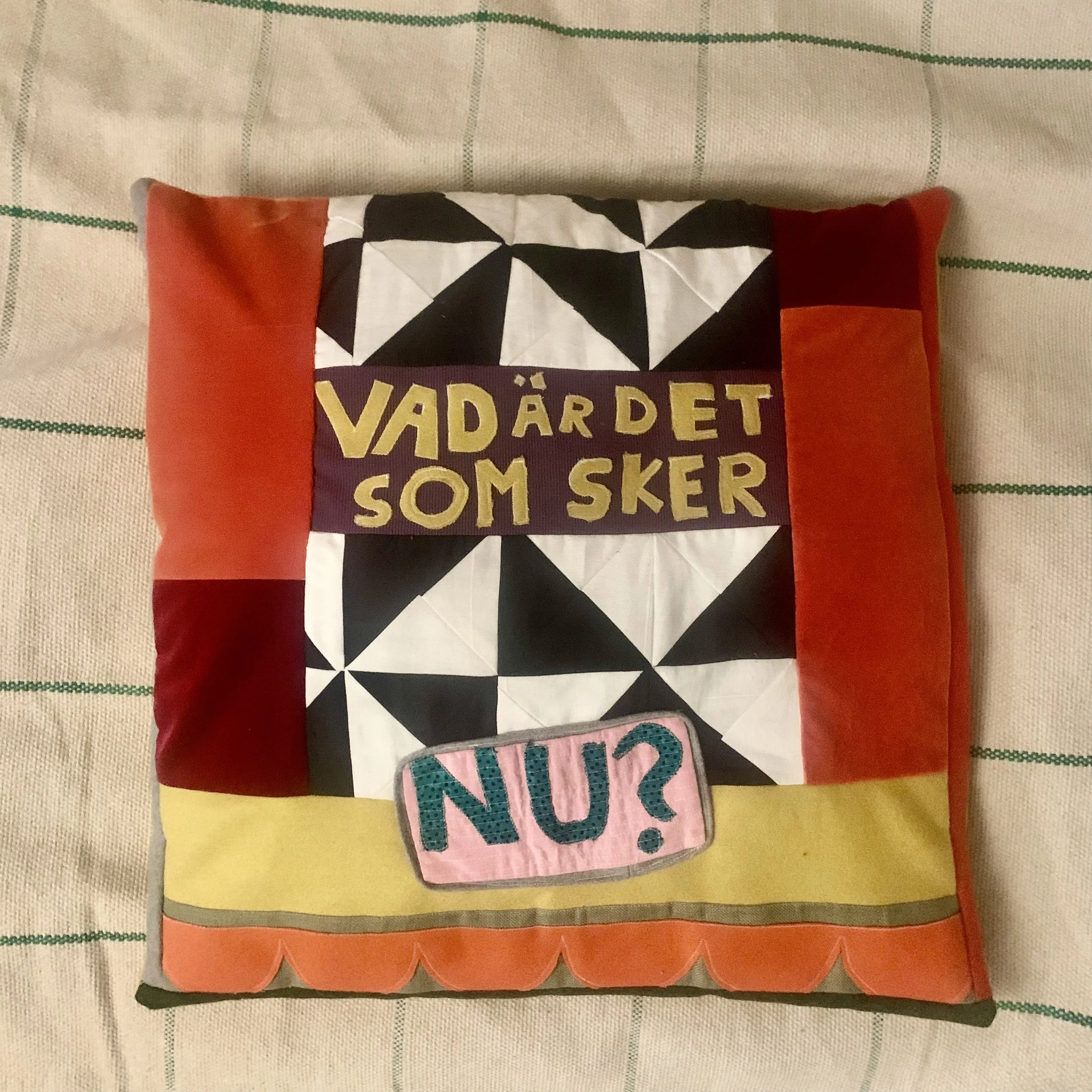 A colorful fabric pillow with a cutout design in red, yellow, and black and white geometric patterns. The pillow has a purple details with yellow Swedish text and a pink patch with teal writing that says "VAD ÄR DET SOM SKER NU?".
