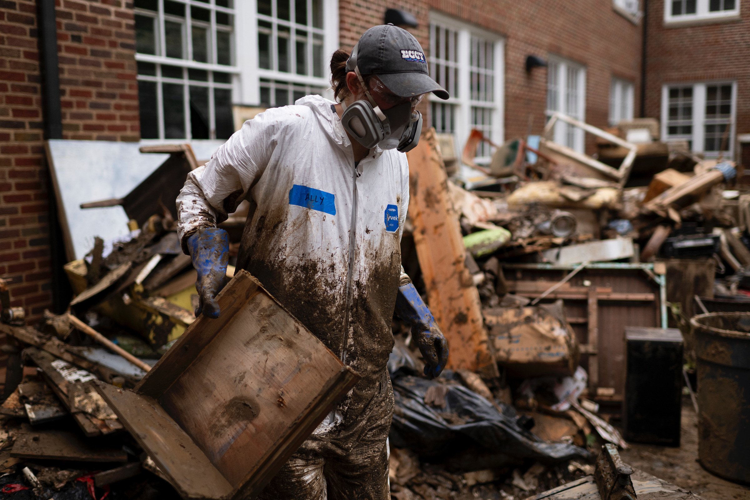 A person wearing a dirty white coveralls labeled 'ALLY', gloves, a cap, and a respirator mask is cleaning up debris in a muddy, chaotic outdoor area filled with broken furniture and trash.