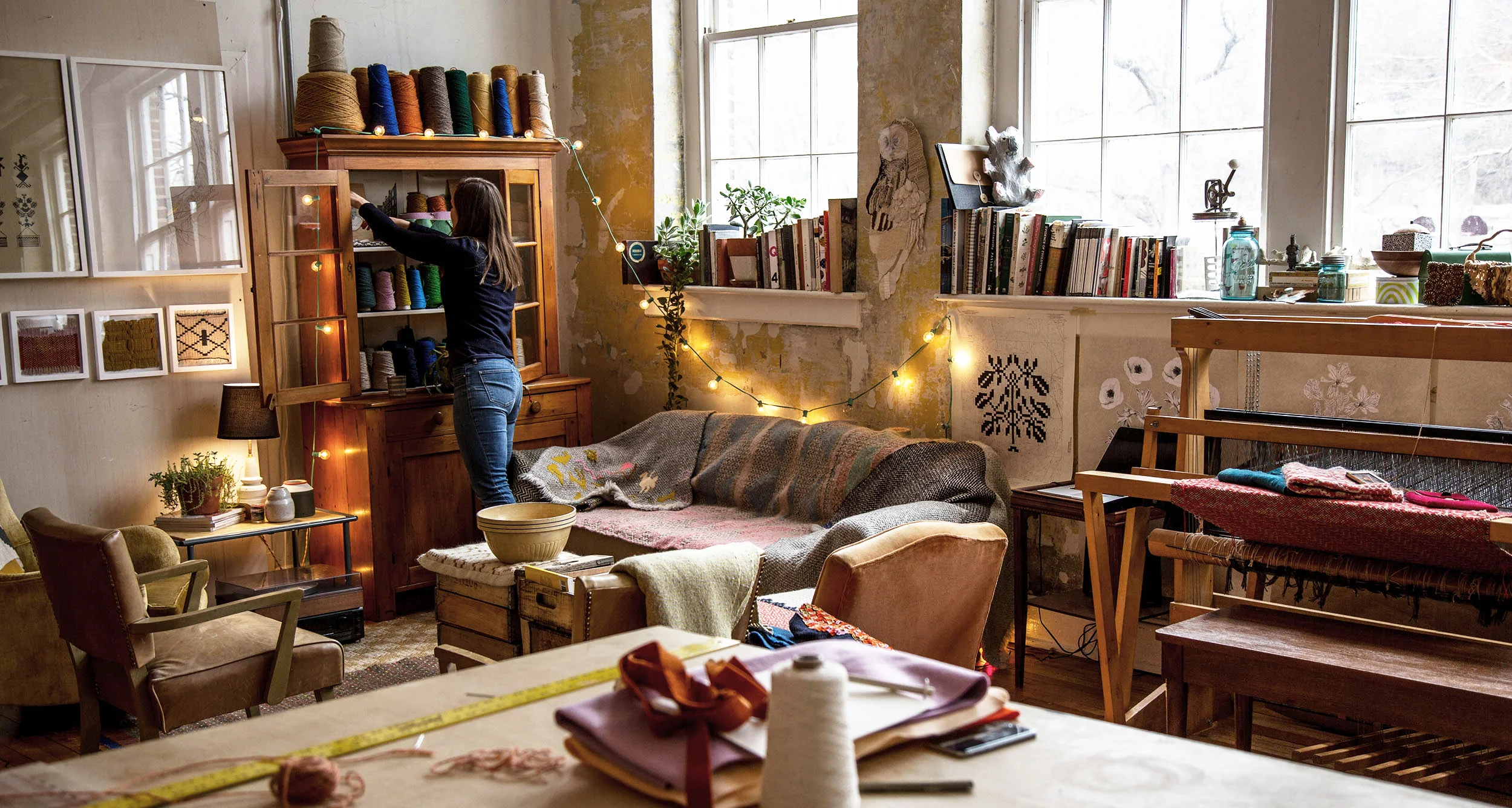 A woman organizing yarn on a wooden cabinet in a cozy, eclectic craft room with large windows, bookshelves, and decorated walls.