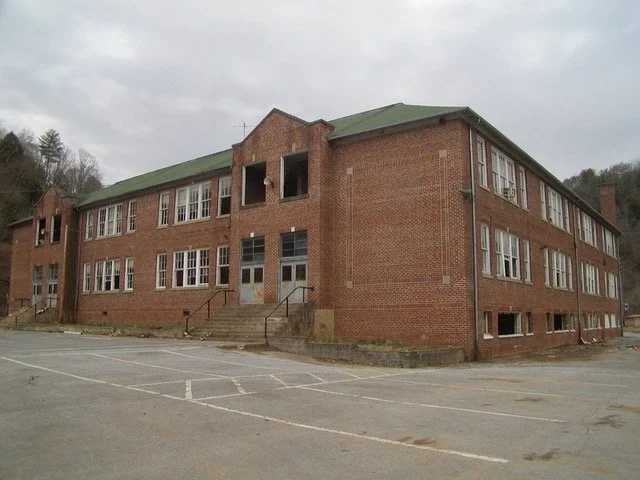 An old brick school building with multiple windows, a green roof, and stairs leading to the entrance, surrounded by an empty parking lot under a cloudy sky.