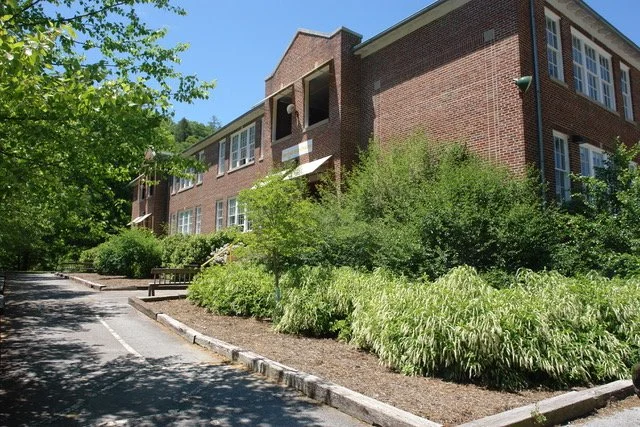 Brick residential building with multiple windows, surrounded by green trees and bushes, with a sidewalk and parked car in front under a blue sky.