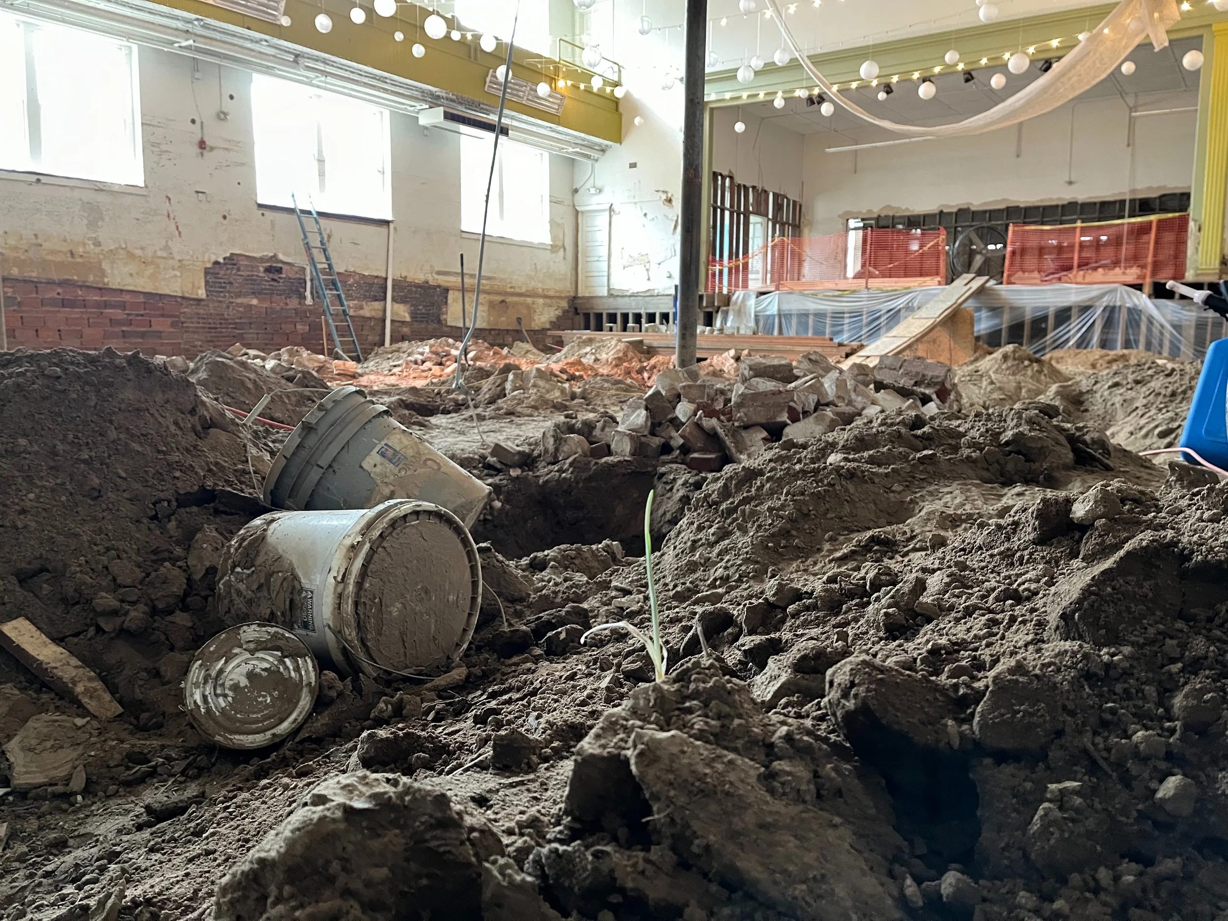 Indoor construction site with dirt, debris, and construction materials, including two buckets, in a partially renovated room with high ceilings, windows, scaffolding, and protective barriers.