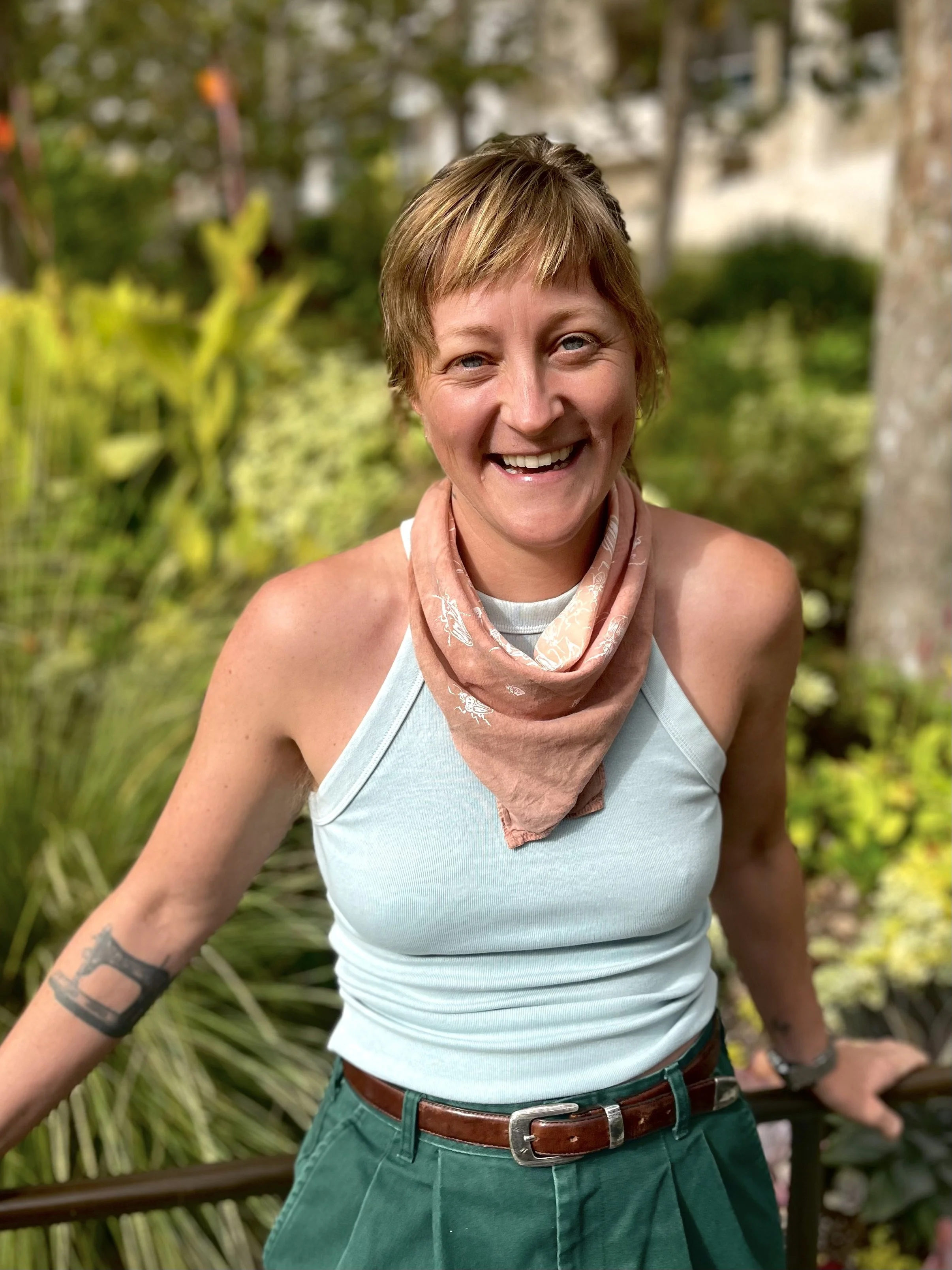 A woman smiling outdoors with short hair, wearing a light blue sleeveless top, a pink bandana, and green pants.