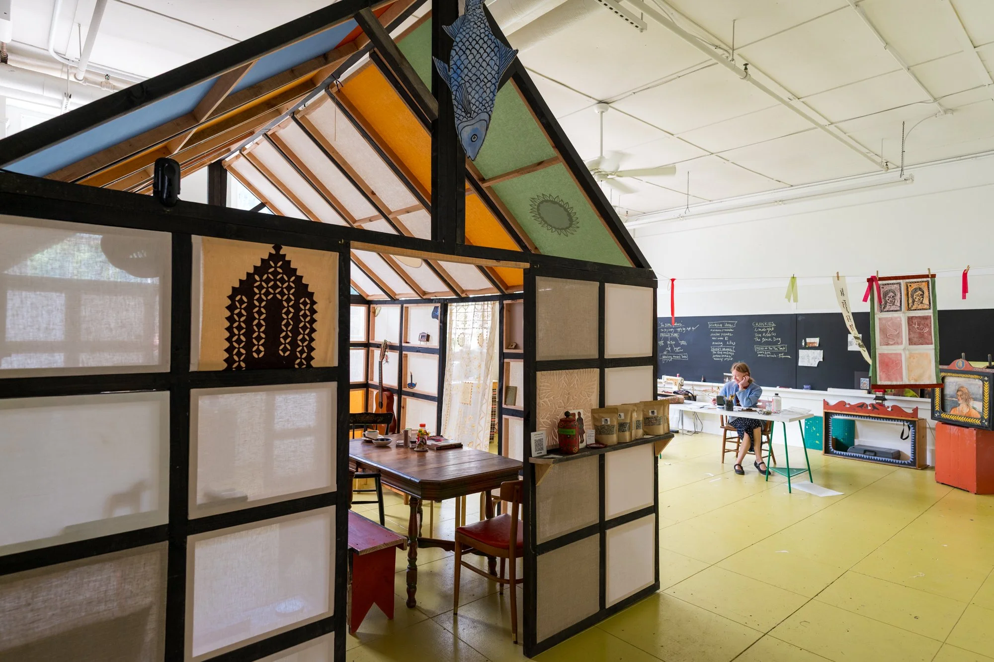 Interior of an art studio with a colorful geometric structure resembling a house, a person working at a desk, and various artwork and decorations on the walls.