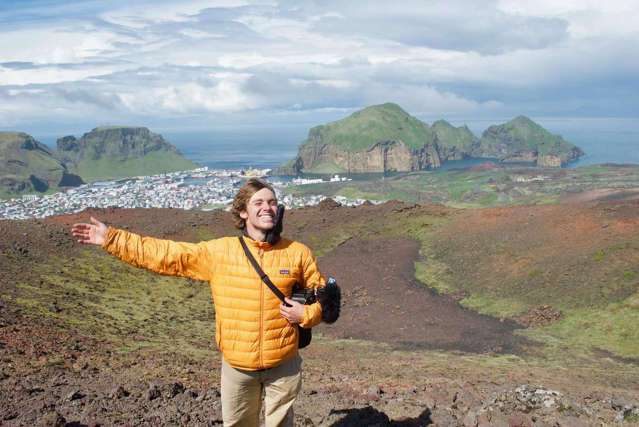 A man in an orange jacket on a mountain trail, smiling with arms outstretched, overlooking a green valley and a coastal town with rocky cliffs and the sea in the background.