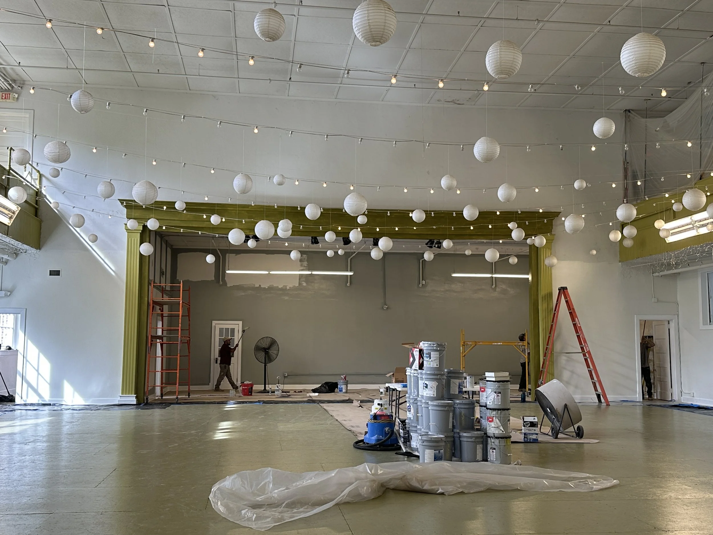 An indoor event space under renovation, with white paper lanterns and string lights hanging from the ceiling, ladders, construction workers, paint buckets, and tools scattered around, and a stage area being prepared.