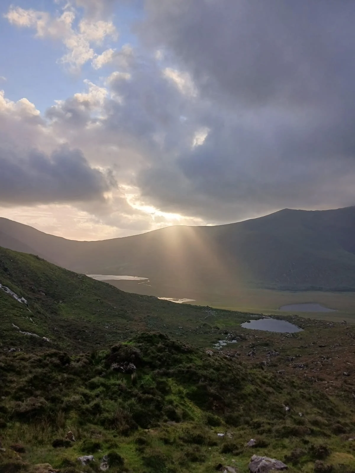 Scenic landscape of rolling green hills and lakes at sunset, under cloudy sky with sunlight breaking through.