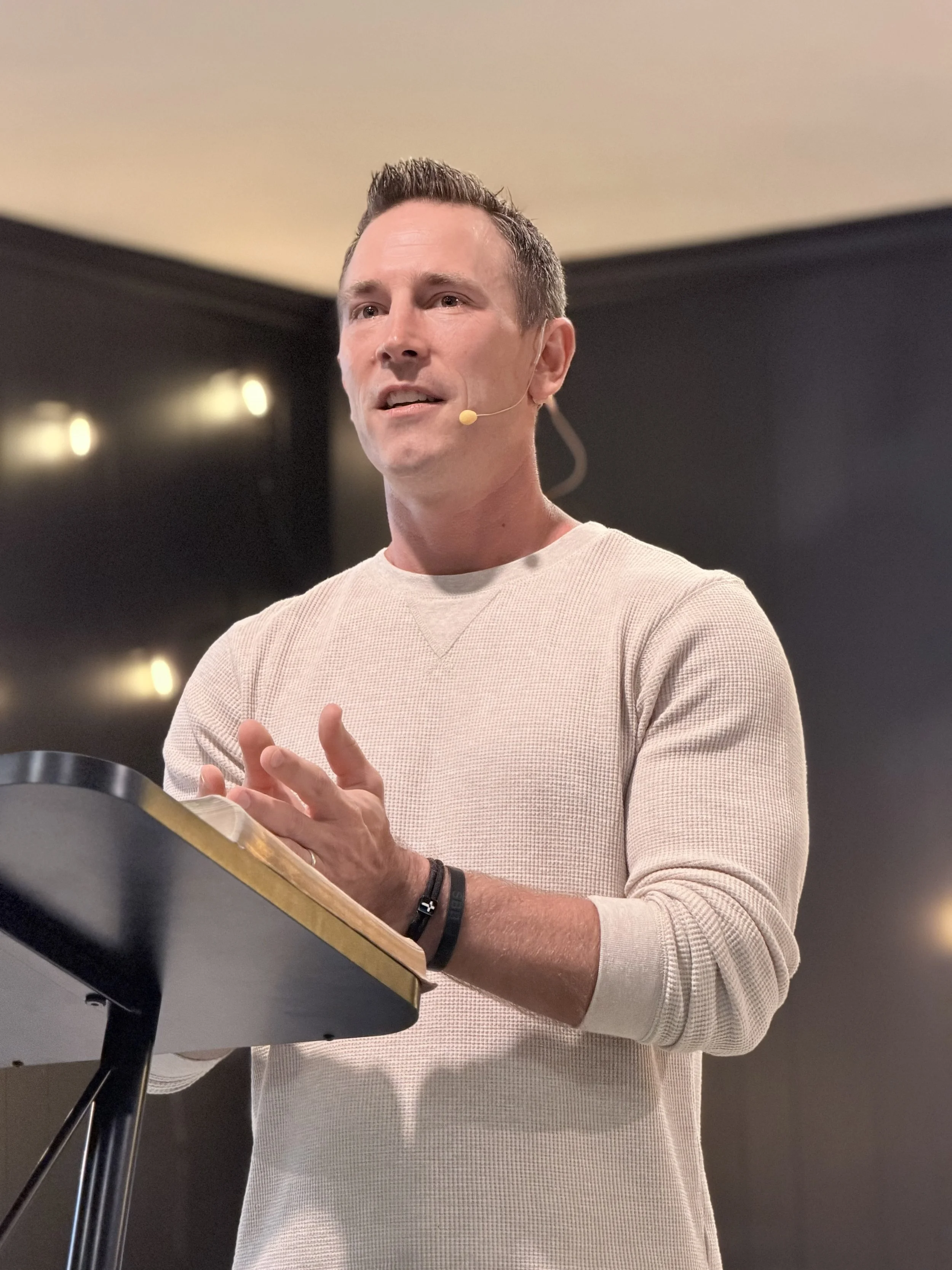 Pastor John Shockley holding a microphone in one hand and a large book in the other, with a large screen behind him displaying graphics and text, including the word 'GIVE' and other icons and words related to environmental sustainability.