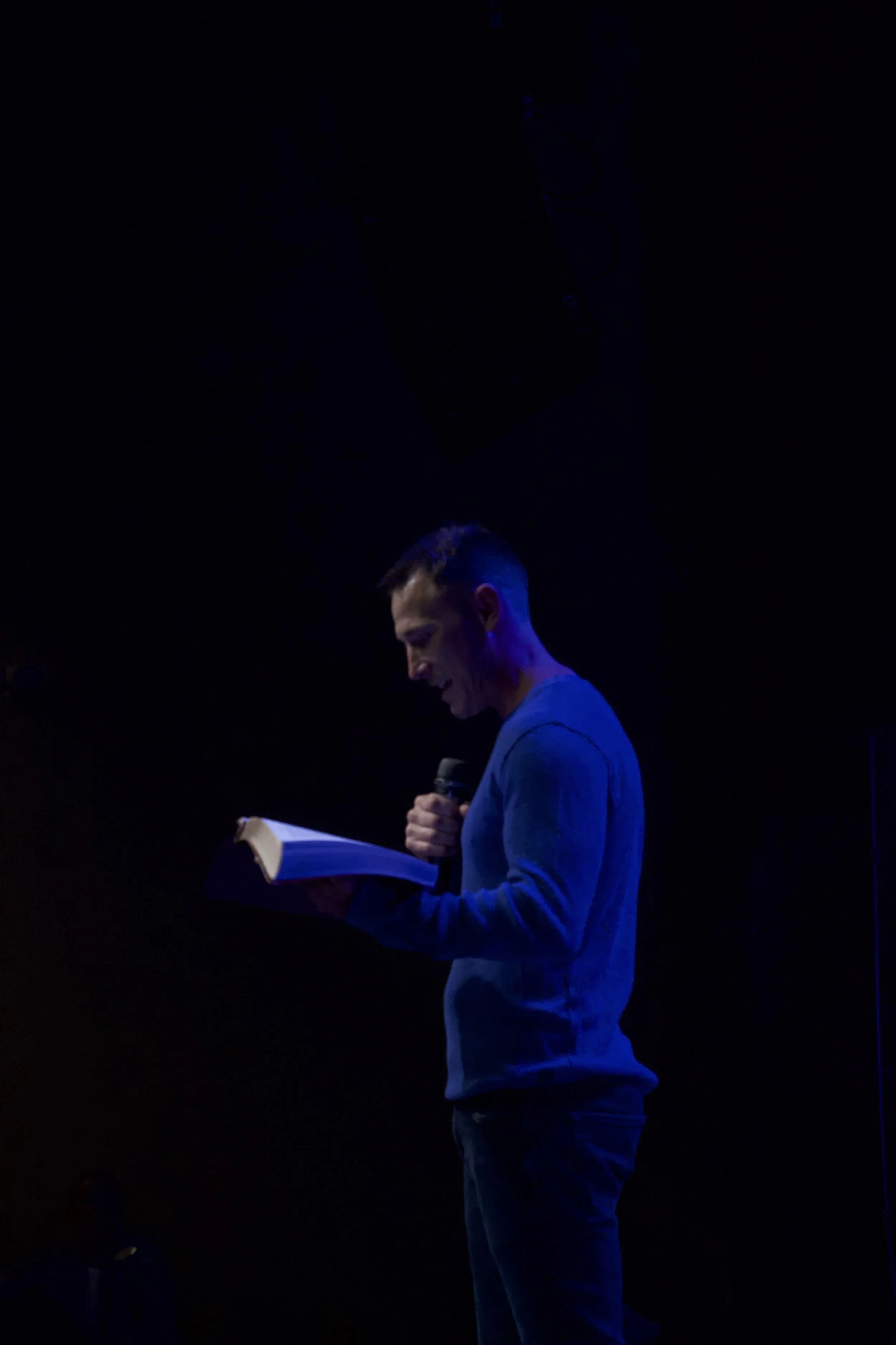Pastor John Shockley holding a microphone in one hand and a large book in the other, with a large screen behind him displaying graphics and text, including the word 'GIVE' and other icons and words related to environmental sustainability.