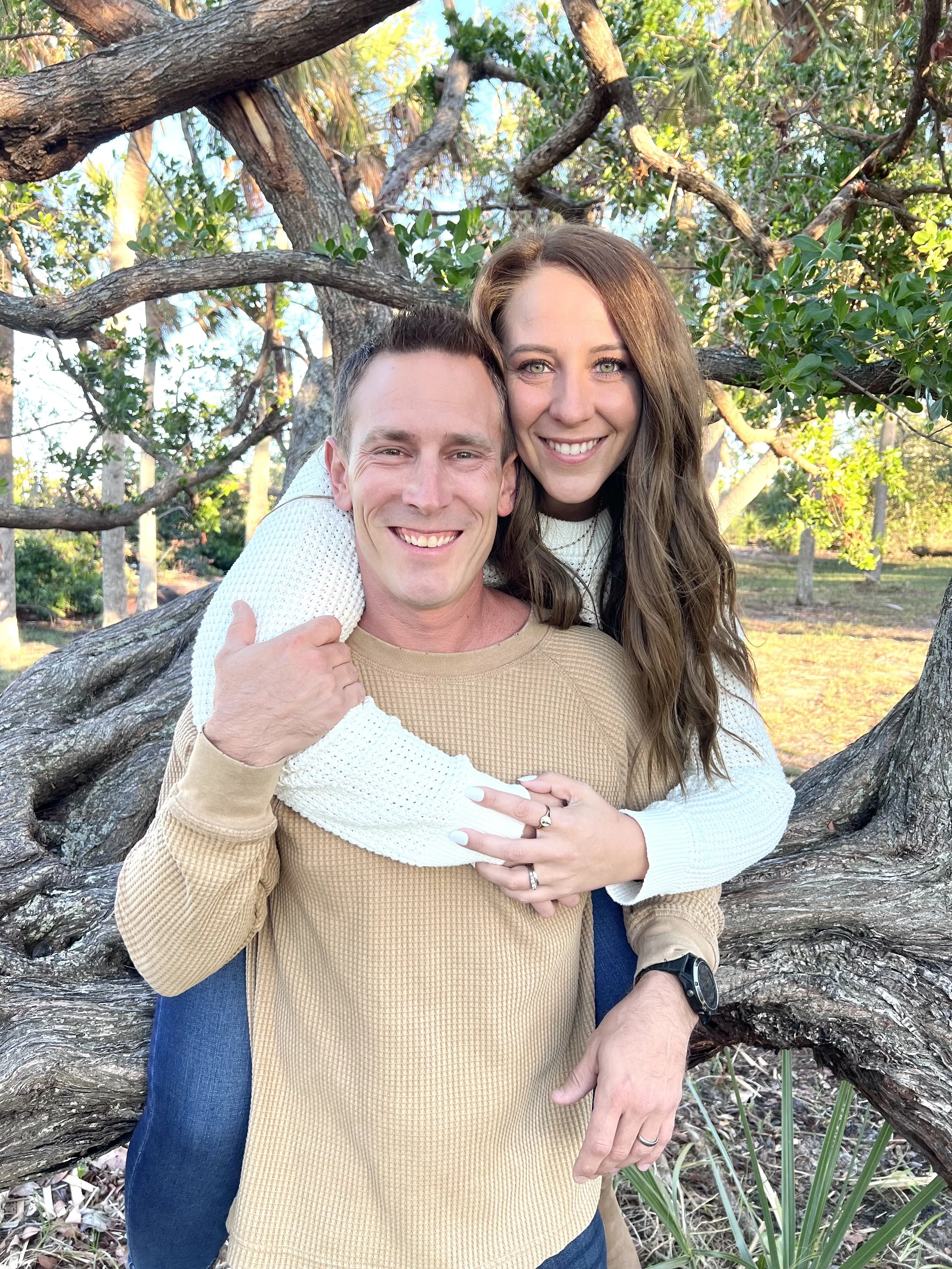 A smiling couple, a man and a woman, outdoors with trees in the background, the woman on the man's back, both looking at the camera.