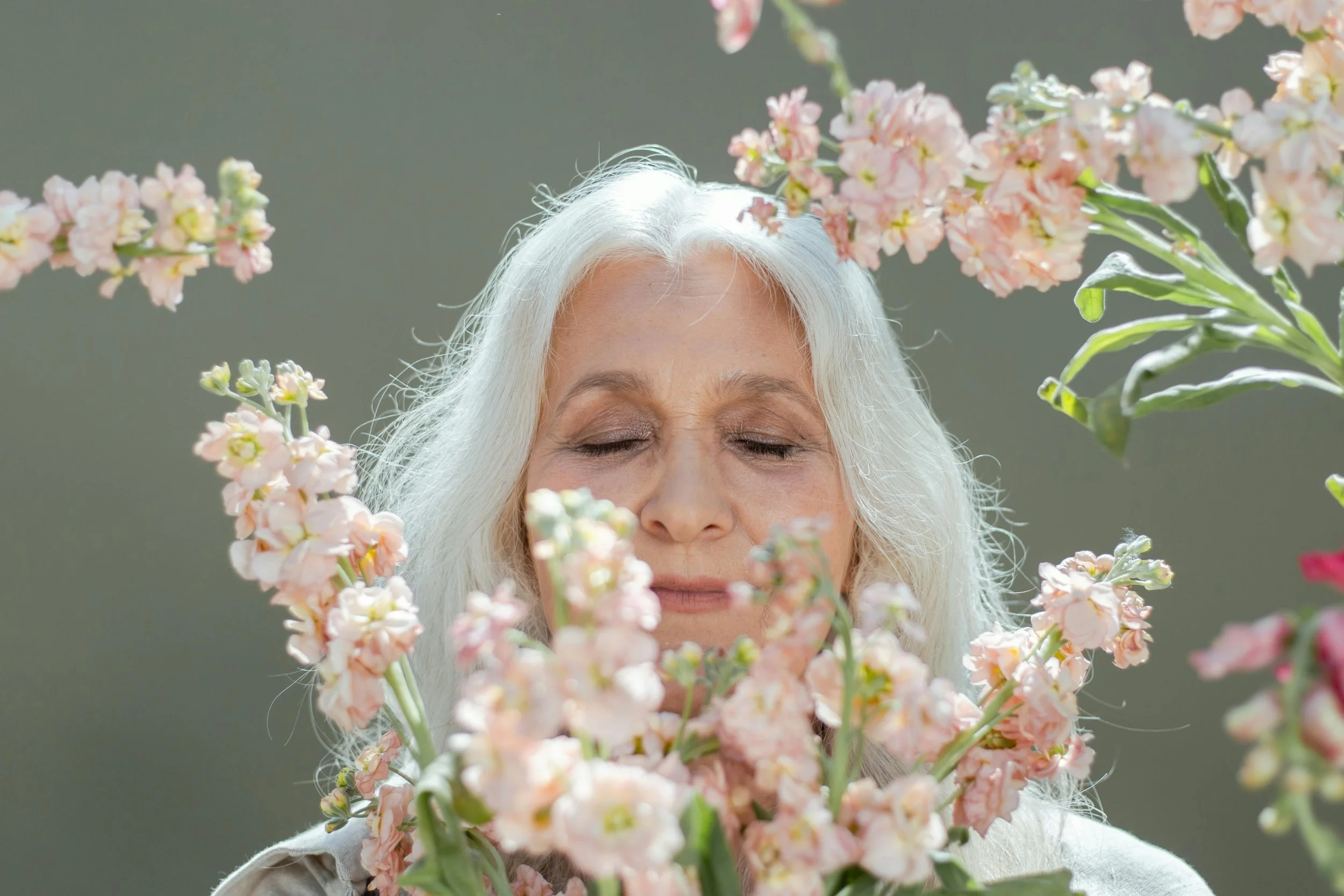 An serene elderly woman with long white hair is surrounded by pink and white flowers.