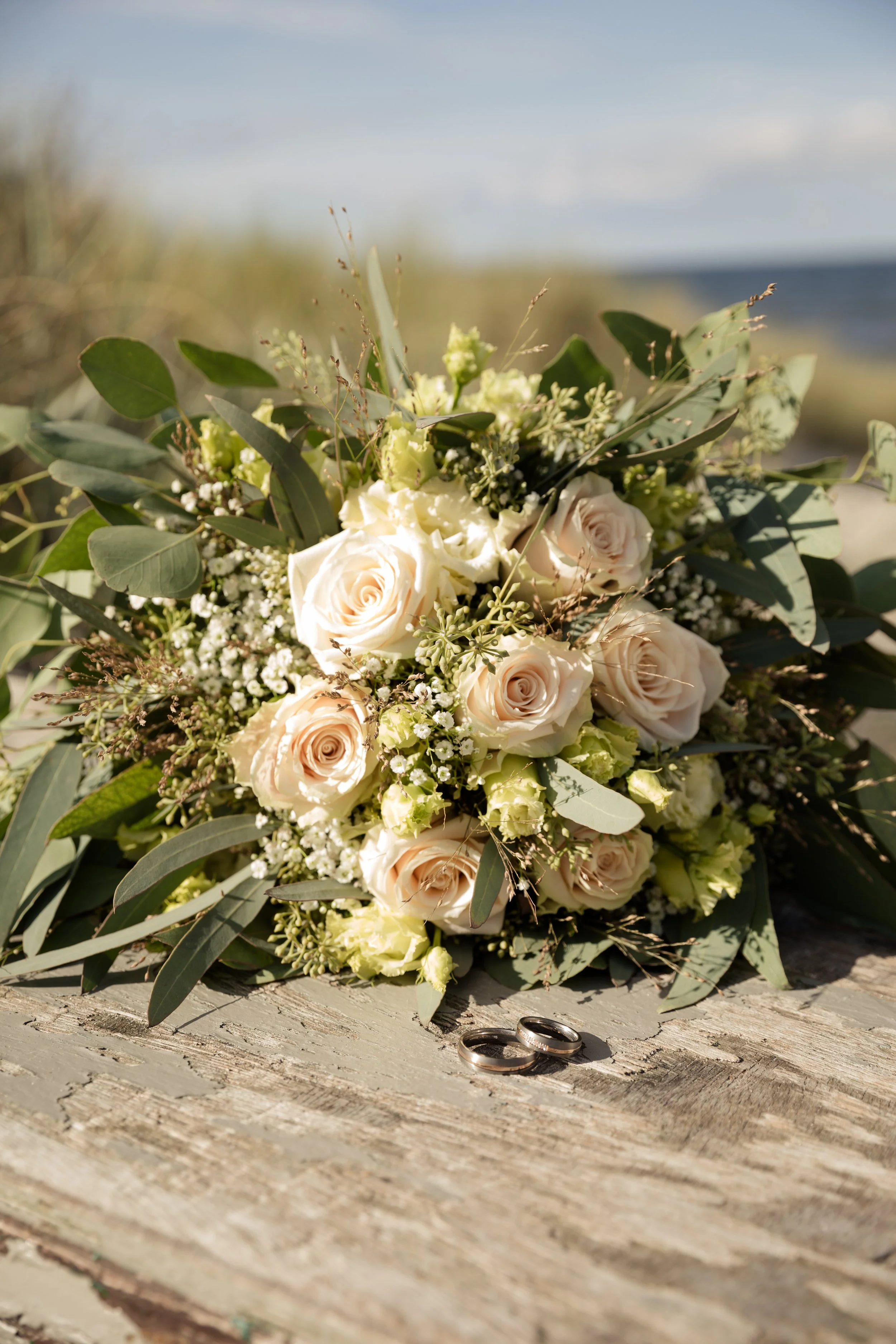 A bouquet of white and cream roses, baby's breath, and eucalyptus leaves rests on a weathered wooden surface, with two wedding rings in front of it, outdoors with a blurred background of sky and water.