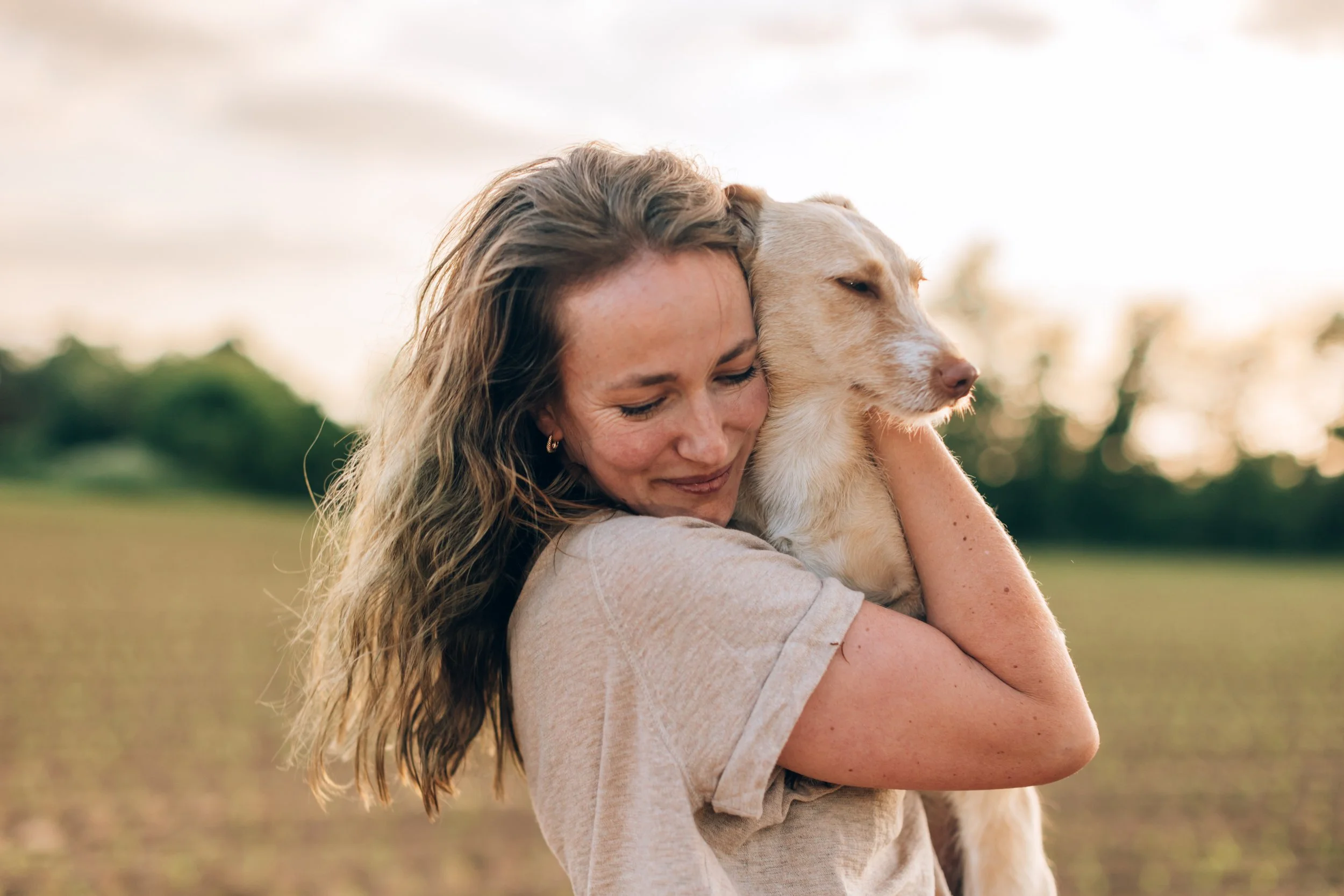 A woman hugging a light-colored dog outdoors during sunset.