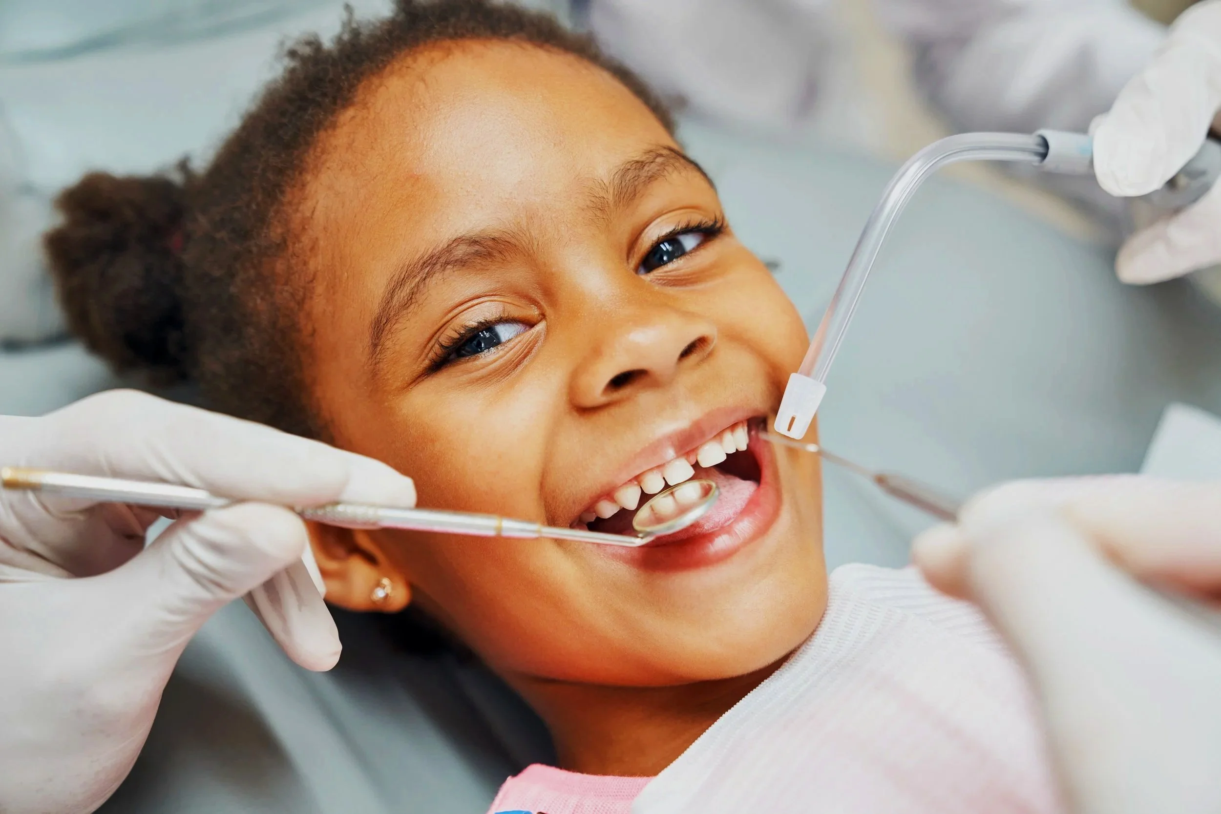 A young girl smiling while at the dentist, undergoing a dental examination with dental tools and suction in her mouth.