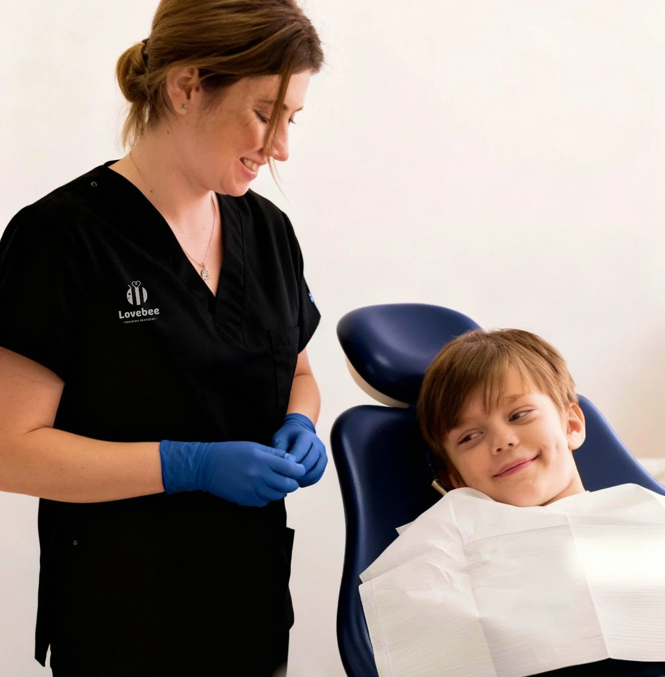 A dental professional smiling at a young boy patient lying in a dental chair, wearing a dentist coat and blue gloves.