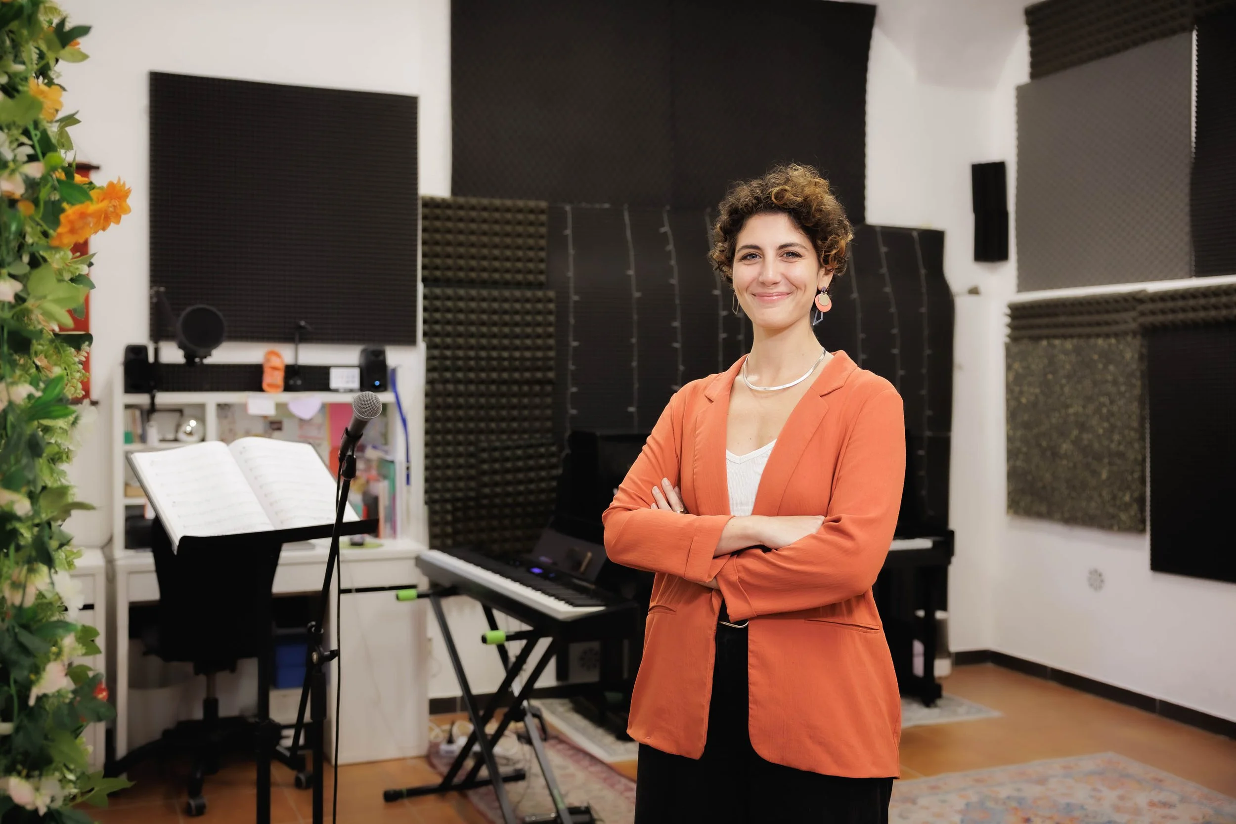 Una donna con capelli riccioli corti, sorridente, indossa una giacca arancione e si trova in uno studio di registrazione con strumenti musicali, microfono e pannelli acustici.
