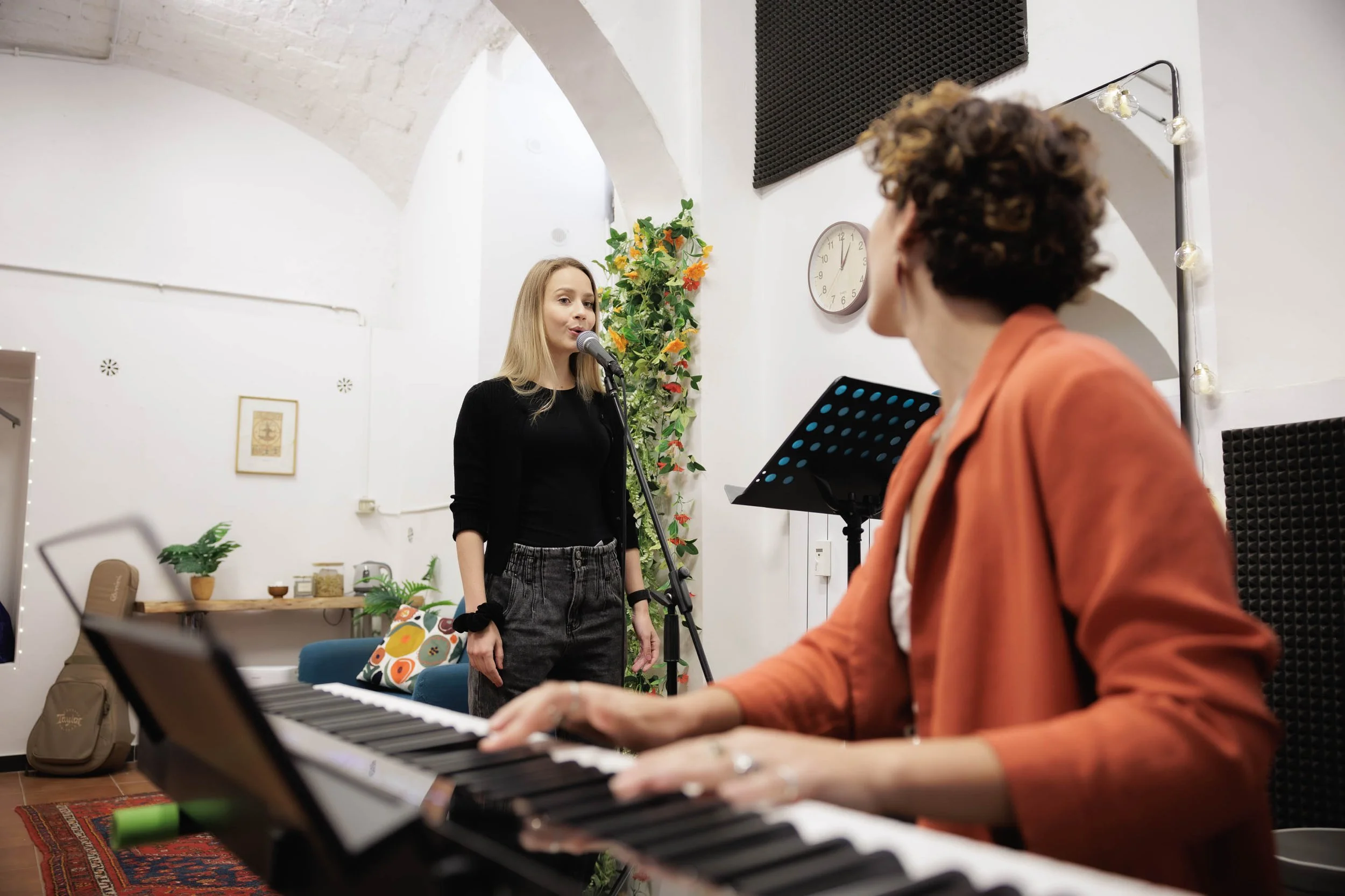 Una donna con cappotto nero canta davanti a un microfono, mentre una persona con giacca arancione suona il pianoforte in un ambiente di musica con decorazioni e luci