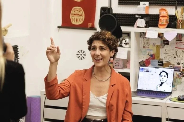 Una donna con capelli ricci, vestita con una maglietta bianca e una giacca arancione, che sta parlando o spiegando qualcosa in un ambiente interno, con uno sfondo di scrivania, computer e vari oggetti.