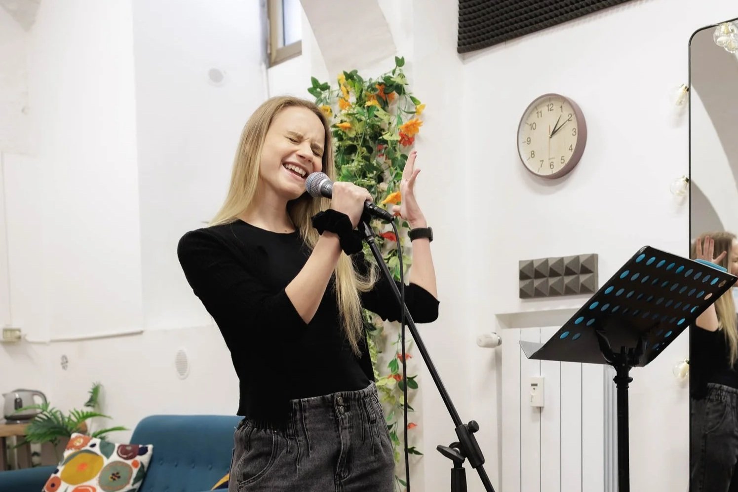 Una ragazza con capelli biondi canta con passione in un luogo chiuso, con un microfono in mano. Sullo sfondo, un orologio e un quadro con sui bassi assi acustici.