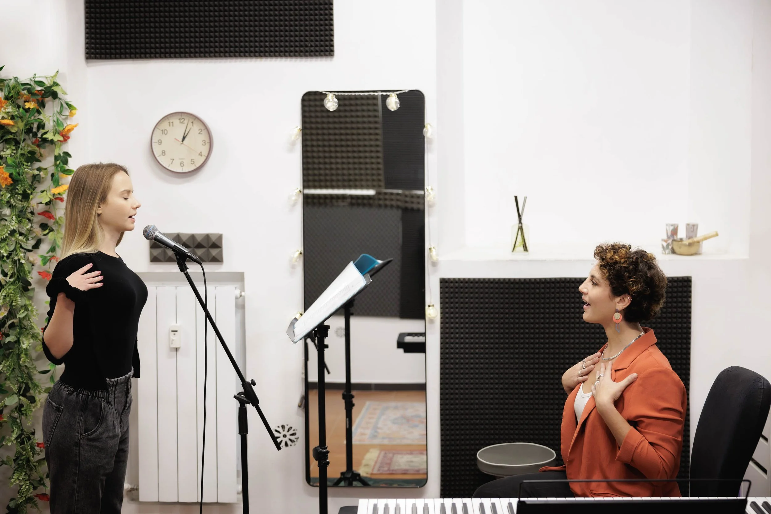 Una ragazza canta di fronte a una donna seduta che la guarda e sorride, in una stanza dedicata alle prove con strumenti musicali e rivestimenti acustici.