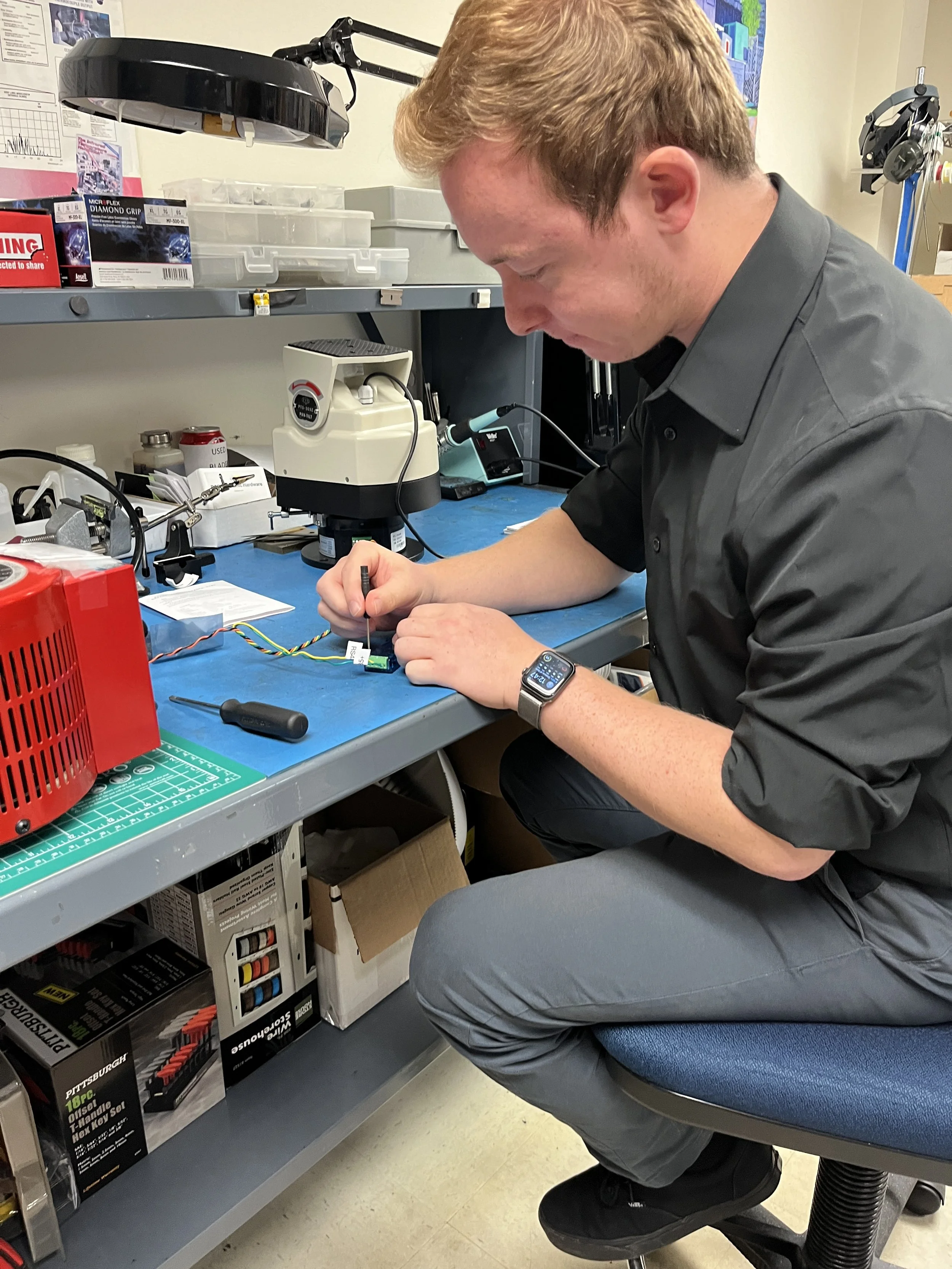 A man working on electronic components at a lab bench with tools, a microscope, and equipment around him.