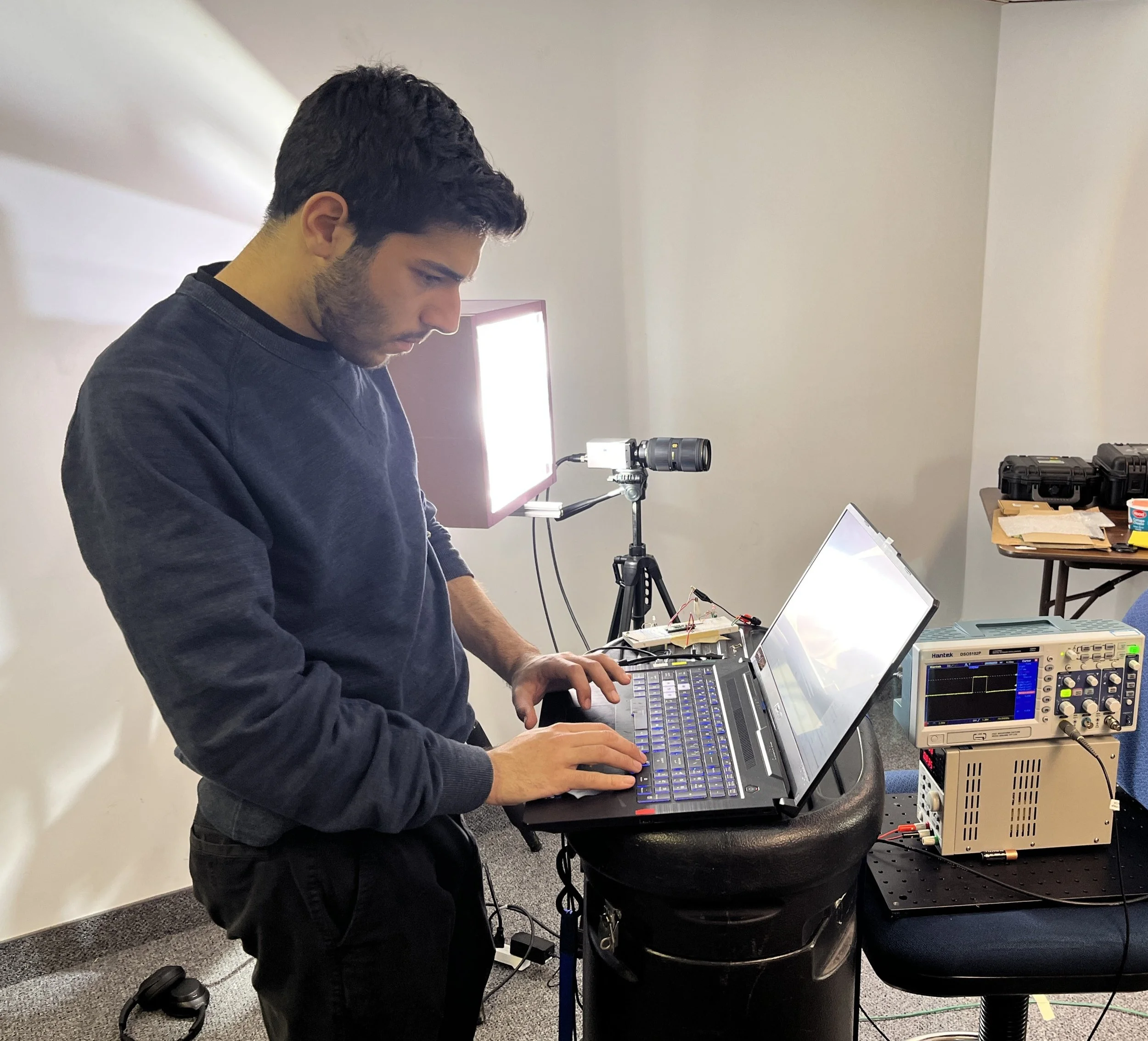 Man working on a laptop connected to electronic testing equipment in a laboratory setting.