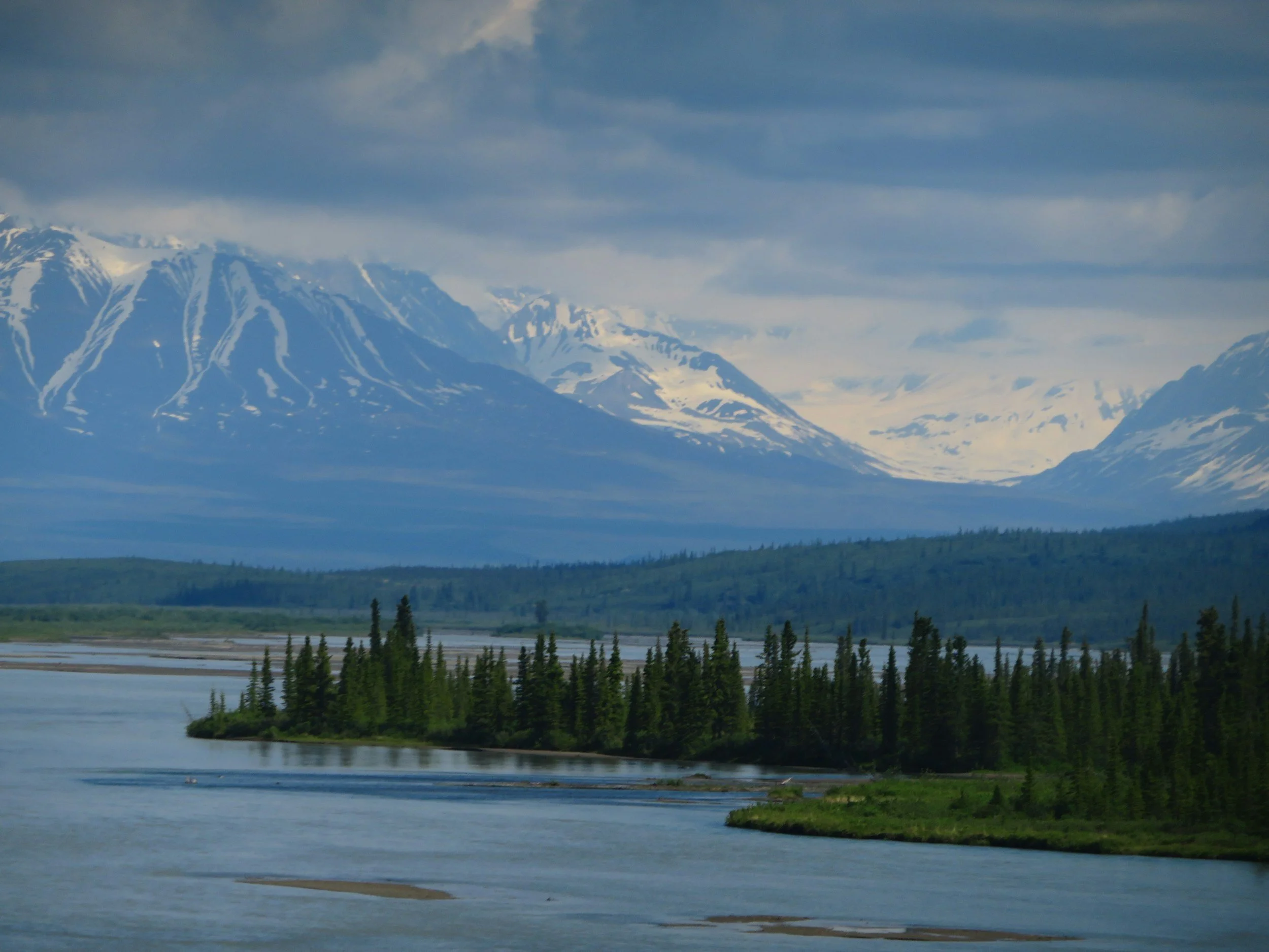 Snow-capped mountains in the background with a lake and green trees in the foreground.