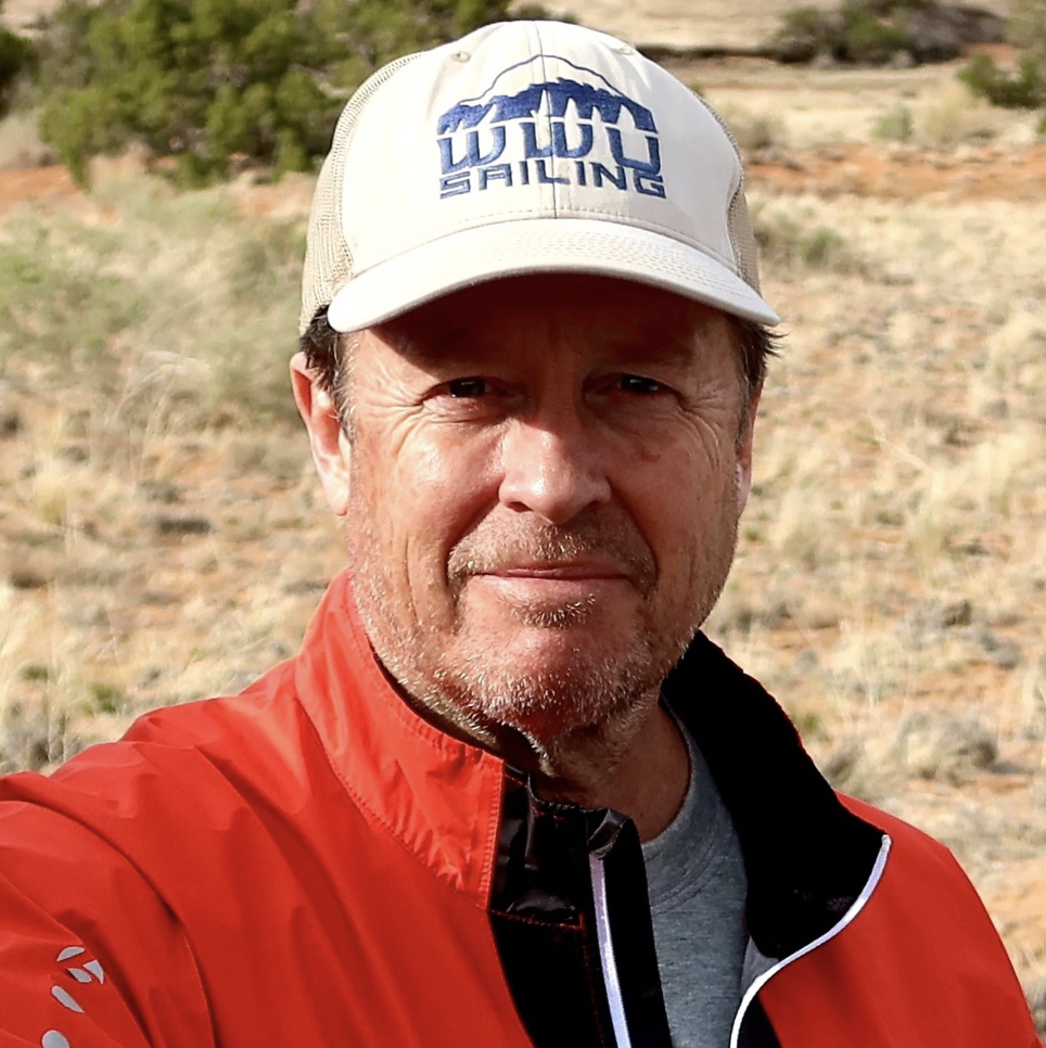A middle-aged man outdoors wearing a beige cap with 'WLOO Sailing' logo, a red jacket, and a gray shirt, with a background of desert-like terrain with rocks and sparse vegetation.