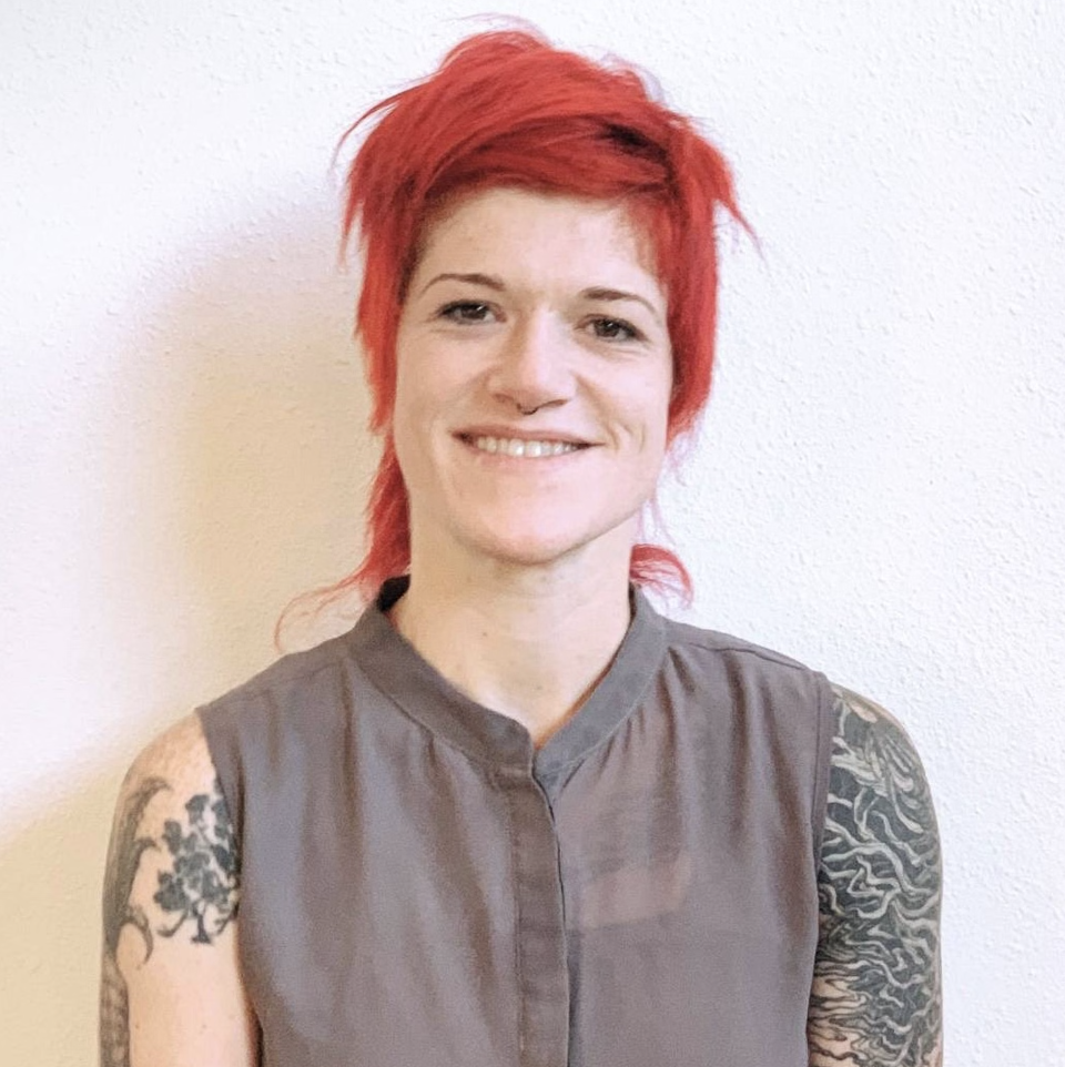 A smiling woman with short, spiky red hair, wearing a sleeveless gray top, standing against a white wall.