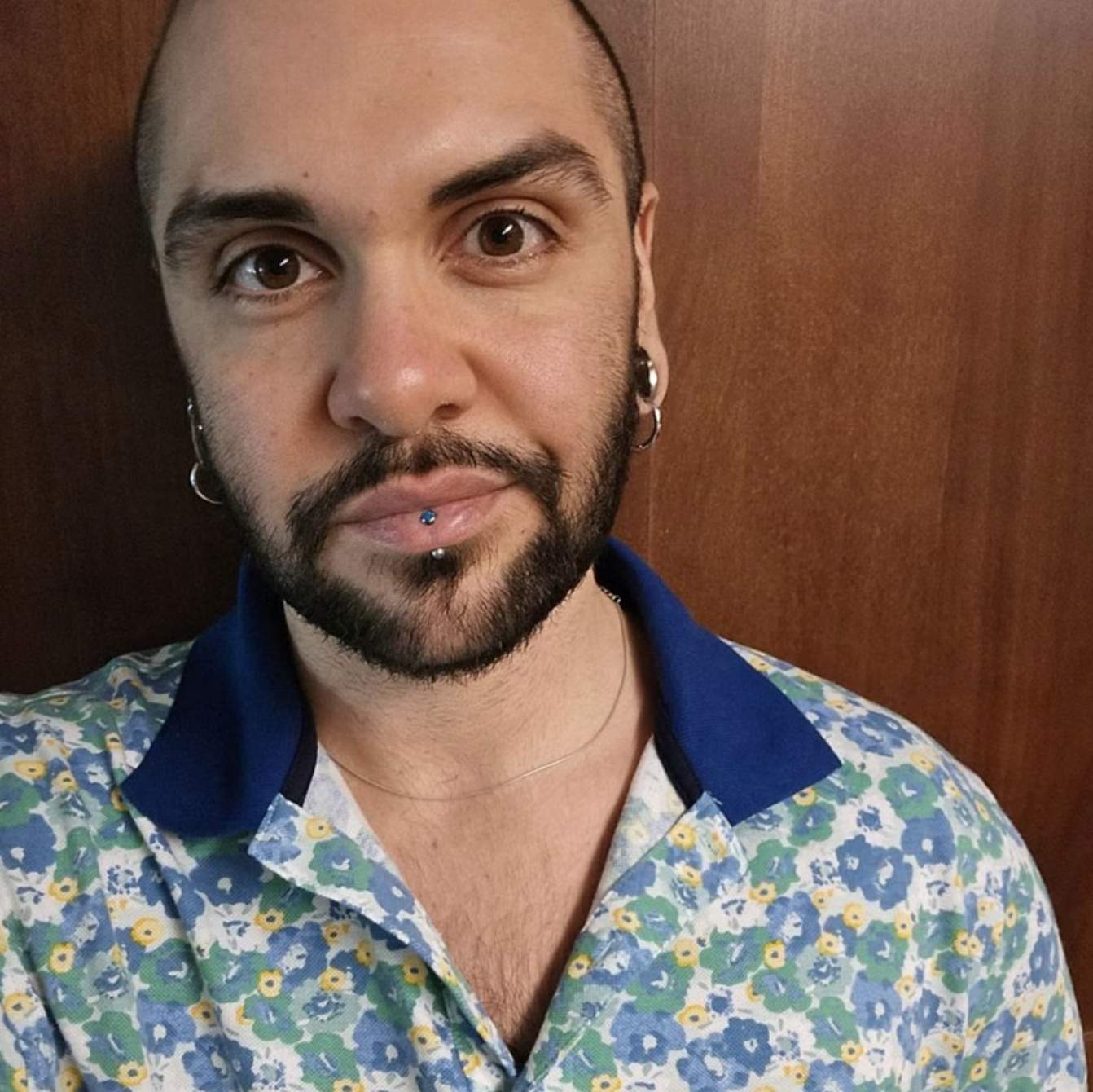 Close-up of a man with tattoos, piercings, and a floral shirt, standing against a wooden background.